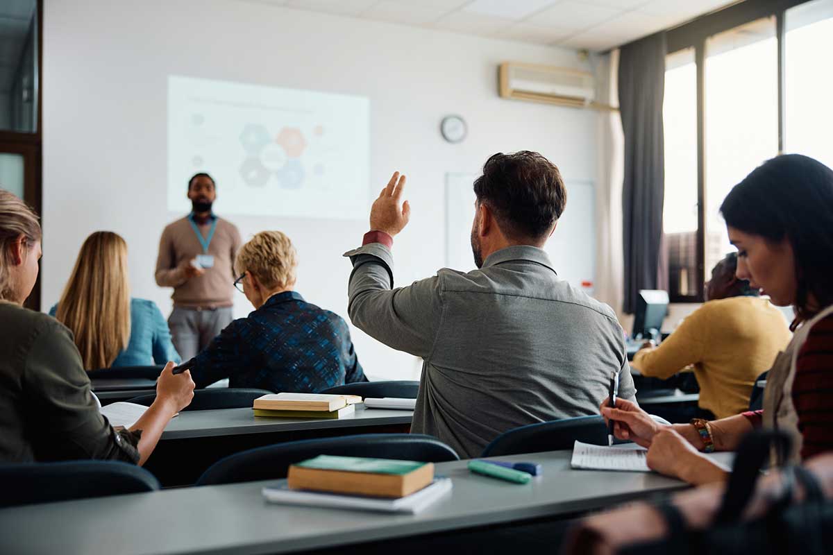 A classroom setting with an instructor at the front and a participant raising their hand to ask a question.