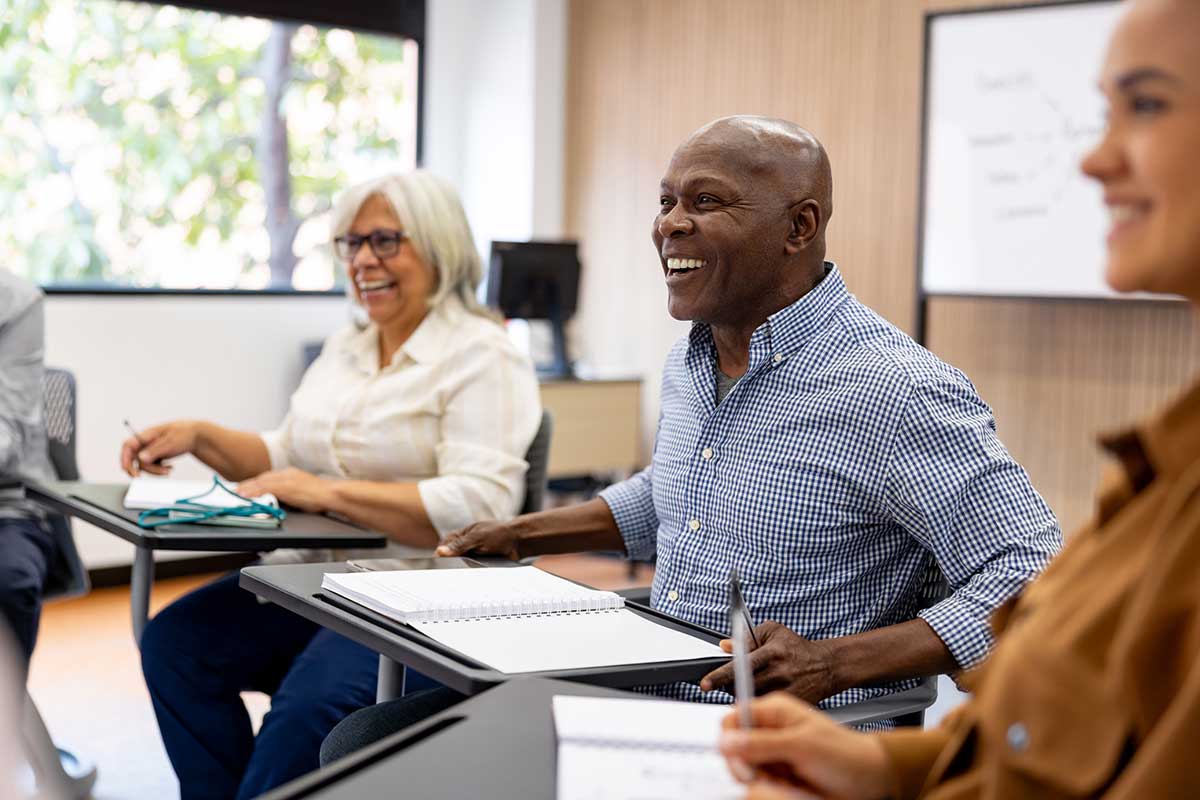 Two older adults sitting at desks, smiling and engaged in a classroom or workshop setting.