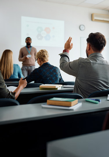 A classroom setting with an instructor at the front and a participant raising their hand to ask a question.
