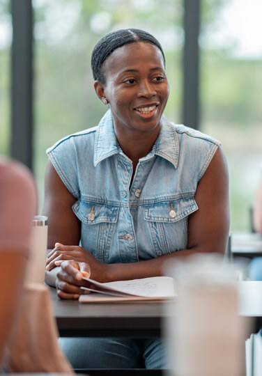 A woman sitting at a table in a group setting, smiling while listening during a discussion.