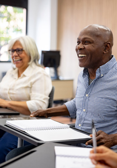 Two older adults sitting at desks, smiling and engaged in a classroom or workshop setting.