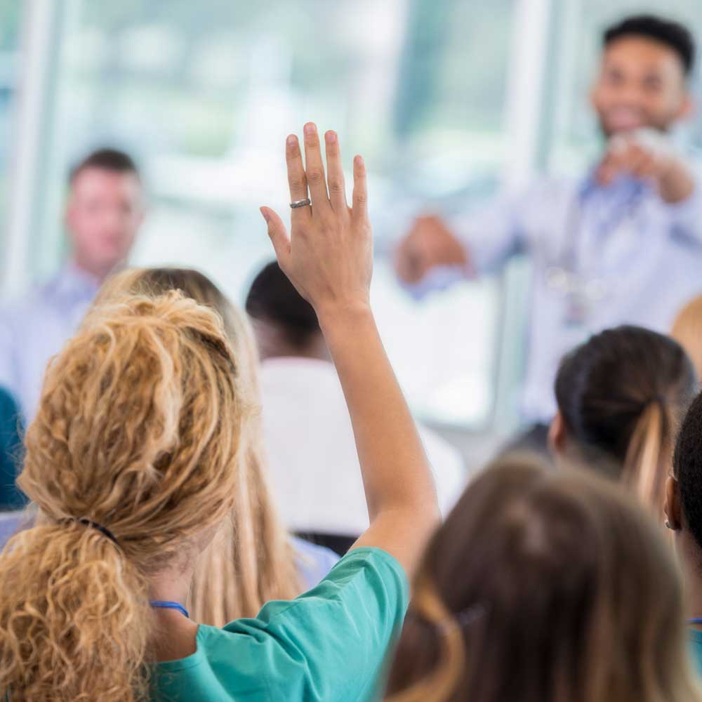 A group setting with a person raising their hand during a presentation while others look toward the speaker.